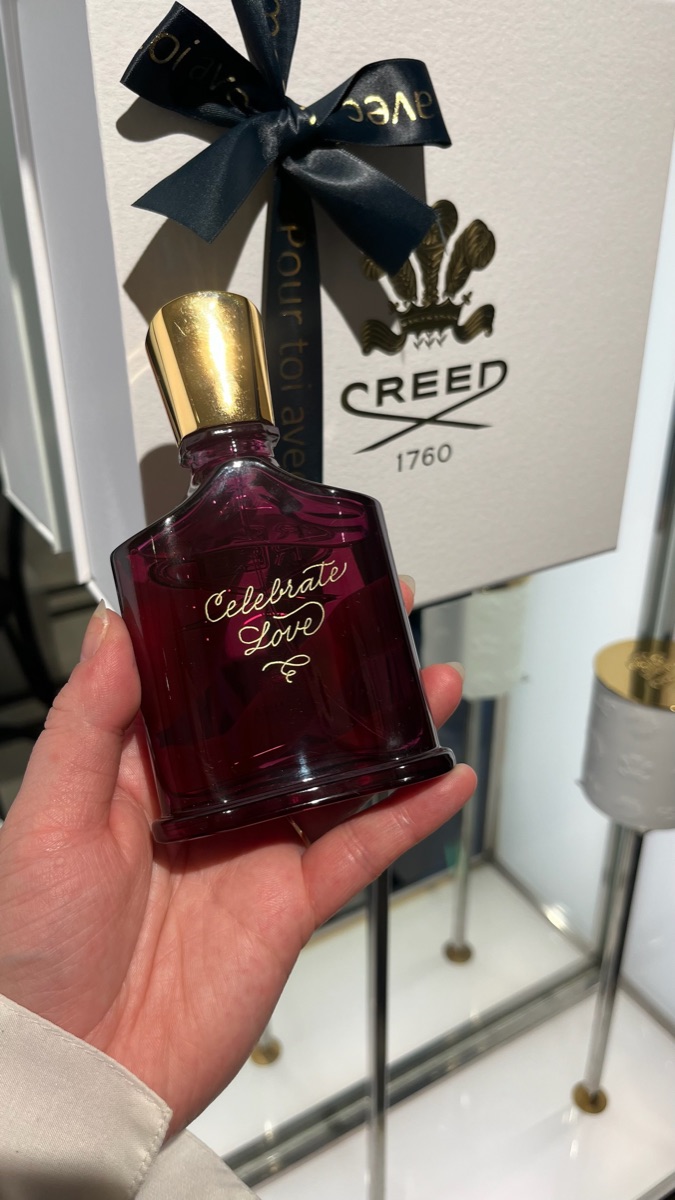 Flacon Creed bordeaux gravé à la main « Celebrate Love » en doré, tenu devant son coffret cadeau noué d'un ruban
