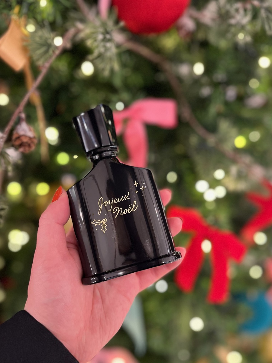Flacon de parfum noir gravé à la main « Joyeux Noël » en doré, avec une petite branche de houx, tenu devant un sapin de Noël illuminé