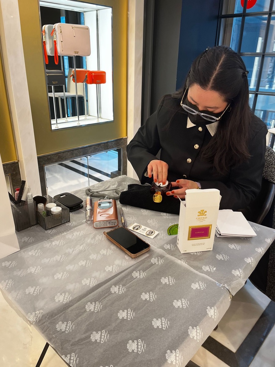 Cathy en train de graver un flacon de parfum à la main sur un stand Creed en boutique, avec ses cires et le carnet des clients