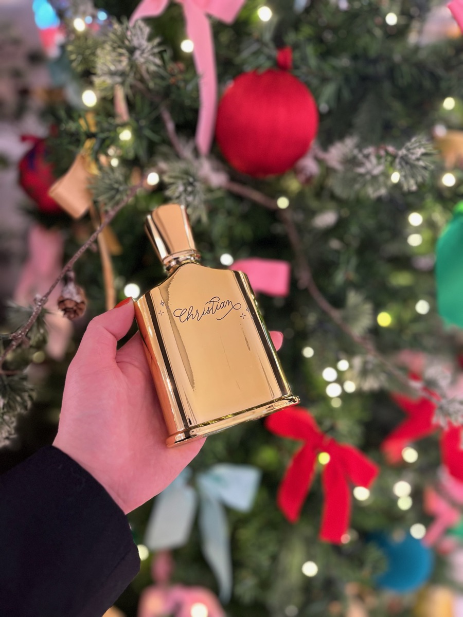 Flacon de parfum doré gravé à la main du prénom « Christian » en lettres cursives, tenu devant un sapin de Noël illuminé