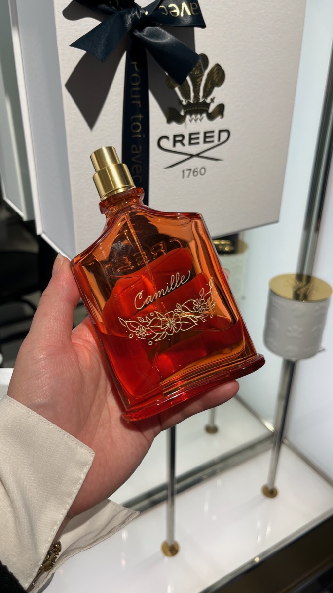 Flacon Creed au verre rouge, prénom « Camille » gravé à la main en doré avec un petit ornement floral, tenu devant son coffret