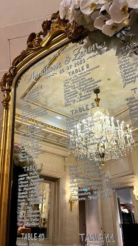 Plan de table de mariage calligraphié à la main en blanc sur un grand miroir doré du Four Seasons George V, avec lustre en cristal et fleurs blanches.