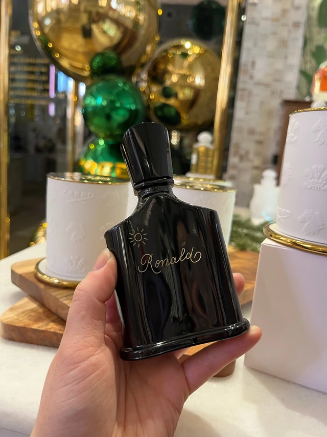 Flacon Creed noir gravé « Ronald » avec un soleil doré, devant des décorations de Noël