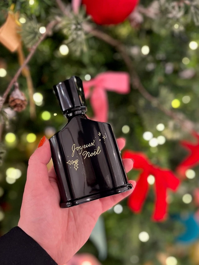 Flacon noir gravé « Joyeux Noël » en doré avec du houx, devant un sapin illuminé