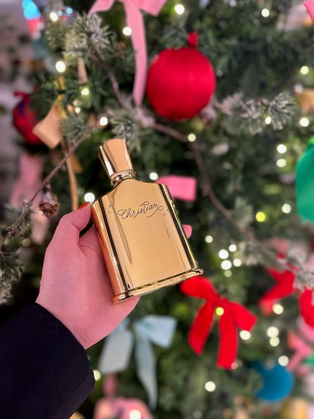 Flacon doré gravé du prénom « Christian » en lettres cursives, devant un sapin de Noël